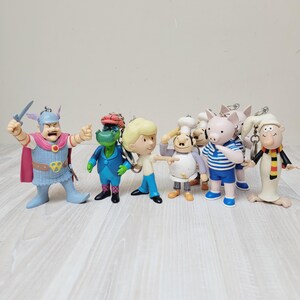 May include: A collection of seven cartoon character keychains featuring a warrior in a winged helmet, a green crocodile in a blue suit, a blonde figure, a chef, a pig in a striped shirt, and a figure in a white robe with a striped scarf.