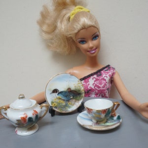 Puede incluir: Un juego de té en miniatura con una tetera blanca, una taza de té y un platillo, y un plato decorativo con un diseño de pájaro. El juego está sobre una superficie gris.