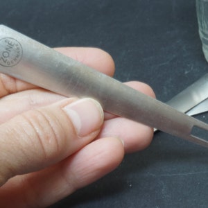 K&ouml;nnte beinhalten: Ein silbernes Metallwerkzeug mit einem Griff und zwei Zinken. Der Griff tr&auml;gt den Aufdruck "ZONE" und "GERMANY".
