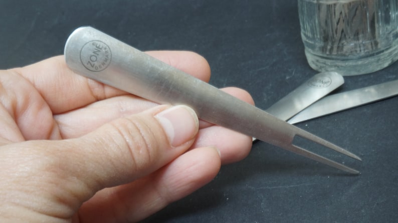 K&ouml;nnte beinhalten: Ein silbernes Metallwerkzeug mit einem Griff und zwei Zinken. Der Griff tr&auml;gt den Aufdruck "ZONE" und "GERMANY".