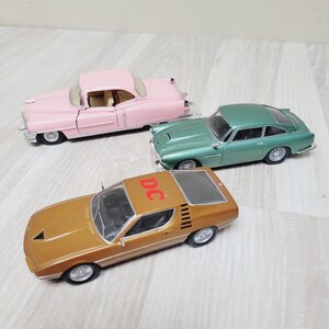 May include: Three die-cast model cars including a pastel pink vintage coupe, a metallic green sports car, and a gold-toned sports car with the letters DC printed in red on the roof. Each vehicle features detailed silver wheels and windows.