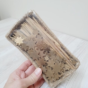 Può includere: Un cestino rettangolare con bordo in juta, riempito con decorazioni a forma di stella in legno e sottili bastoncini di legno. Le stelle sono di varie dimensioni e tonalità di marrone. Il cestino è avvolto in plastica trasparente.