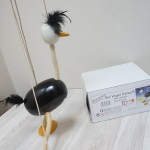 May include: A black and white wooden puppet of an ostrich with a long neck and a black feather on its head. The puppet is on a white wooden table with a white box that says "Der Vogel Strauß" on it.