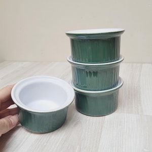 May include: Four green ceramic ramekins with white interiors. The ramekins are stacked in a pyramid shape, with the top ramekin slightly offset from the center.