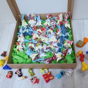 Può includere: Una scatola di cartone marrone piena di piccoli regali avvolti in colori vivaci. La scatola è circondata da una varietà di piccoli giocattoli di plastica colorati.