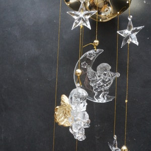 May include: A crystal mobile with a crescent moon, stars, and angels hanging from a gold chain. The moon has a cherub playing a musical instrument.