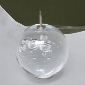 May include: A clear glass sphere with a silver top and many small air bubbles inside. The sphere is sitting on a white surface.