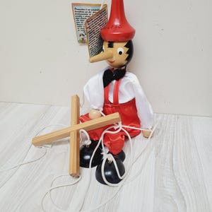 May include: Wooden Pinocchio marionette with a red hat, red trousers, and a white shirt. The puppet has a long nose, black hair, and a black bow tie. The marionette is attached to a wooden cross-shaped control.