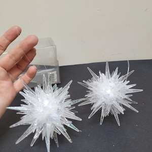 May include: Two clear, star-shaped ornaments with pointed edges and iridescent glitter accents. Each ornament has a small light inside and is suspended by a thin, clear string. The ornaments are on a dark surface.