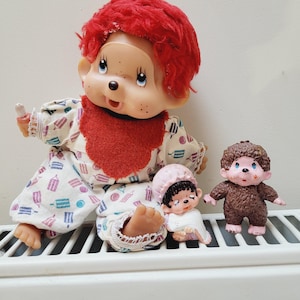 May include: A group of vintage Monchhichi dolls. The main doll has red hair, a red bib, and a patterned outfit. Two smaller dolls are also visible, one brown and one wearing a pink hat and white outfit. The dolls are displayed on a white surface.