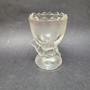 Puede incluir: Huevera de cristal transparente con superficie texturizada y base decorativa. El borde de la taza tiene un diseño en zigzag. La huevera está diseñada para contener un solo huevo.