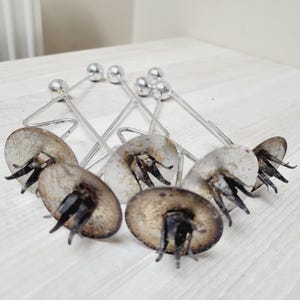 May include: Six silver candle holders with a round base and three prongs. The holders are made of metal and have a silver finish. The prongs are designed to hold candles securely.