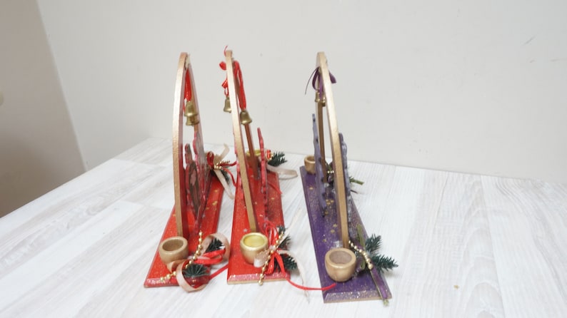 K&ouml;nnte beinhalten: Drei h&ouml;lzerne Weihnachts-Kerzenhalter mit goldenen Akzenten. Die Halter sind mit rotem, violettem und goldenem Glitzer verziert. Jeder Halter hat eine kleine Glocke und einen Kerzenhalter.
