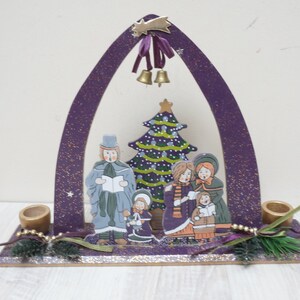 K&ouml;nnte beinhalten: Lilafarbener Weihnachtsbogen aus Holz mit einer bemalten Szene von S&auml;ngern und einem Weihnachtsbaum. Der Bogen hat zwei Kerzenhalter auf jeder Seite.