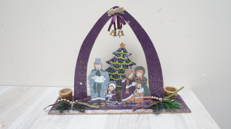 K&ouml;nnte beinhalten: Lilafarbener Weihnachtsbogen aus Holz mit einer bemalten Szene von S&auml;ngern und einem Weihnachtsbaum. Der Bogen hat zwei Kerzenhalter auf jeder Seite.