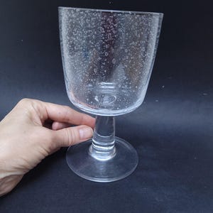 May include: A clear glass goblet with a textured surface, featuring small air bubbles throughout the glass. The goblet has a tall, slender stem and a wide, round base.