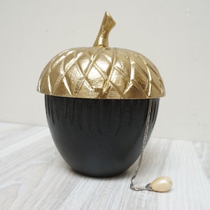 May include: A black and gold acorn-shaped trinket box with a lid. A silver chain with a pearl hangs next to the box.