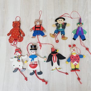 May include: A collection of eight wooden jumping jack ornaments featuring a red bear with gold stars, a brown bear, a child in clogs, a clown, a snowman with a scarf, a snowman with a top hat, a penguin, and a ladybug, all with red pull strings.