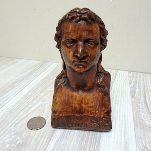 May include: A wooden bust of a man with a serious expression. The bust is brown and has a nameplate that reads "SCHILLER".