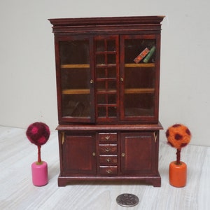 Może przedstawiać: Miniaturowa drewniana biblioteczka z przeszklonymi drzwiami i szufladami. Biblioteczka jest ciemnobrązowa i ma tradycyjny design. Jest ozdobiona dwoma małymi, kolorowymi drzewami z filcu.