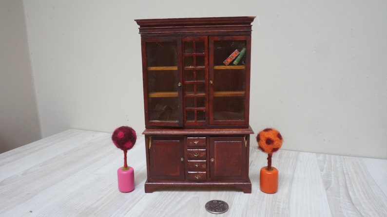 Może przedstawiać: Miniaturowa drewniana biblioteczka z przeszklonymi drzwiami i szufladami. Biblioteczka jest ciemnobrązowa i ma tradycyjny design. Jest ozdobiona dwoma małymi, kolorowymi drzewami z filcu.