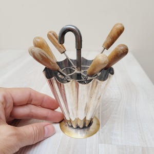 Könnte beinhalten: Eine silberfarbene, geriffelte Servierschale mit goldfarbenem Sockel, die sechs Spieße mit Holzgriff enthält. Die Schale hat einen gebogenen Griff. Die Spieße sind so angeordnet, dass sie einem Regenschirm ähneln.