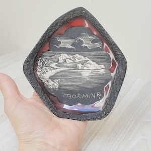 May include: A black and red ceramic tile with a white etched image of a mountain landscape and the word "Taormina".