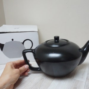 May include: A black ceramic teapot with a lid and a handle. The teapot is sitting on a white wooden table.