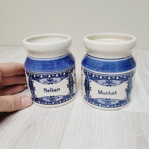 Könnte beinhalten: Zwei Keramik-Gewürzgläser mit blau-weißem Design. Die Gläser haben eine zylindrische Form mit weißem Rand und Boden. Die Gläser tragen die Wörter "Nelken" und "Muskat" in einem dekorativen Rahmen.
