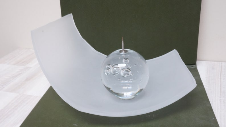 May include: A clear glass sphere with air bubbles inside, resting on a white curved glass base.