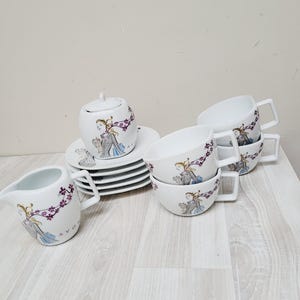 Puede incluir: Juego de té de porcelana blanca con azucarero con tapa, jarrita para la leche, platillos y tazas. Cada pieza está decorada con una mujer con un vestido azul y detalles florales morados. La jarrita para la leche tiene la palabra "AVON".