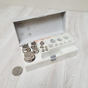 May include: A vintage white box containing a set of metal weights of various sizes, used for precision measurements. The weights are arranged in a compartmentalized tray. A silver coin is next to the box. The box is on a light wood surface.