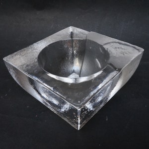 Op de afbeelding: Een asbak van helder glas met een vierkante basis en een rond, komvormig interieur.