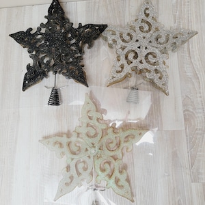 Op de afbeelding: Drie decoratieve stervormige kerstboomtoppers. De sterren zijn gemaakt van een glinsterend materiaal en hebben een delicaat, ingewikkeld ontwerp. De sterren zijn zwart, zilver en goud.