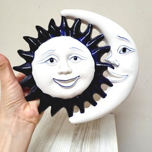 Könnte beinhalten: Keramische Wanddekoration mit einer weißen Sonne und einem Mond mit blauen Akzenten. Die Sonne hat ein lächelndes Gesicht und der Mond hat ein einzelnes Auge.