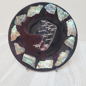 Può includere: Un piatto decorativo nero con accenti iridescenti di conchiglia di abalone. Il piatto presenta una mappa della Nuova Zelanda al centro, con i nomi delle città. Il piatto è esposto su un supporto trasparente.