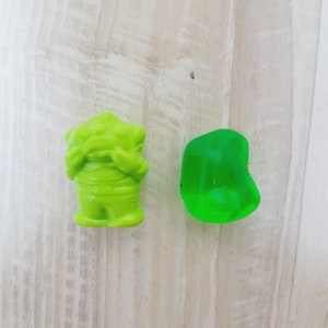 Op de afbeelding: Twee groene gummy snoepjes, een in de vorm van een monster en de andere een eenvoudige rechthoek, op een lichte houten achtergrond.
