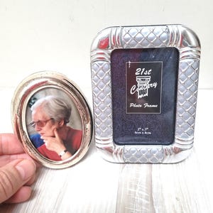 Puede incluir: Dos marcos de fotos plateados. El marco más grande es rectangular con un patrón acolchado y el texto "21st Century Photo Frame 2" x 3" 6cm x 8cm". El marco más pequeño es ovalado y tiene una superficie lisa.