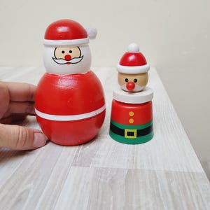 Op de afbeelding: Twee houten kerstversieringen in de vorm van de kerstman. De ene is een grote, afgeronde figuur met een rode body, witte afwerking en een kerstmannengezicht. De andere is kleiner, met een rode top, een witte basis en een groene onderkant met een zwarte riem.
