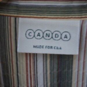 Puede incluir: Una etiqueta blanca con el texto "CANDA" en c&iacute;rculos negros y el texto "MADE FOR C&A" en letras negras. La etiqueta est&aacute; unida a una camisa a rayas con rayas blancas, beige, marrones y negras.