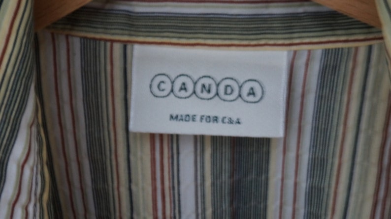 Puede incluir: Una etiqueta blanca con el texto "CANDA" en c&iacute;rculos negros y el texto "MADE FOR C&A" en letras negras. La etiqueta est&aacute; unida a una camisa a rayas con rayas blancas, beige, marrones y negras.