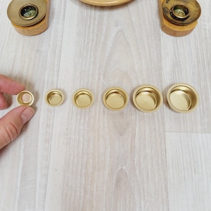 May include: A collection of small, gold-colored metal bowls of varying sizes, arranged in a row. A hand holds one of the smallest bowls. The bowls are on a light wood surface. In the background, a wooden decorative piece with small figurines is visible.