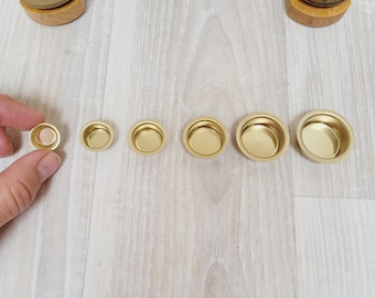Può includere: Una collezione di piccole ciotole in metallo dorato di varie dimensioni, disposte in fila. Una mano tiene una delle ciotole più piccole. Le ciotole sono su una superficie di legno chiaro. Sullo sfondo, un pezzo decorativo in legno con piccole statuine.