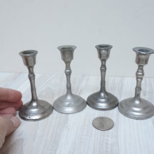 以下が含まれることがあります： シンプルなデザインの銀色の金属製のキャンドルホルダーが4つ。キャンドルホルダーは白い木製の表面に一列に並んでいます。