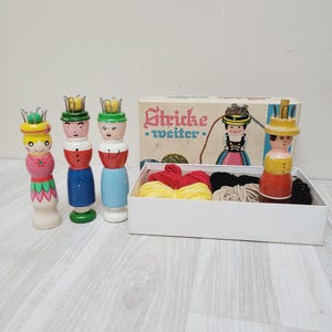 Könnte beinhalten: Ein Vintage-Strickset mit einer Box mit der Aufschrift "Stricke weiter" und vier Holzsteckpuppen. Die Puppen sind in leuchtenden Farben mit kronenförmigen Aufsätzen bemalt. Die Box enthält Garn in Rot, Gelb, Schwarz und Beige.