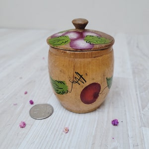 Może przedstawiać: Mały drewniany słoik z pokrywką, pomalowany wzorem czerwonych i fioletowych owoców z zielonymi liśćmi. Słoik stoi na białym drewnianym blacie.