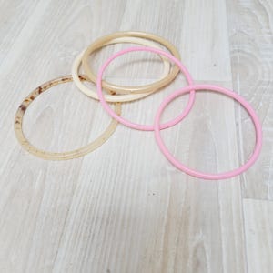 May include: A selection of circular bangles in different colours. The bangles include light pink, cream, tan, and marbled brown. They are made of a smooth, glossy material and are arranged on a light wood-grain surface.