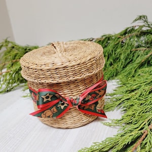 May include: A round, woven basket with a lid, tied with a festive ribbon. The ribbon is green with red accents and teddy bear and star patterns. The basket is sitting on a white wooden surface, with green foliage in the background.