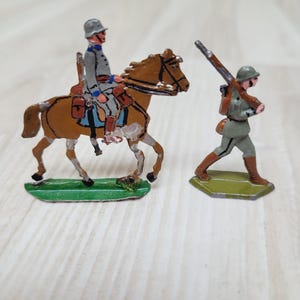 Puede incluir: Dos soldados de juguete vintage. Uno es un soldado montado a caballo marrón, y el otro es un soldado que lleva un rifle. Ambas figuras están pintadas con detalles y se apoyan en bases verdes. Las figuras probablemente son de metal.