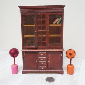 Puede incluir: Un gabinete de madera en miniatura con puertas de vidrio y cajones. El gabinete es de color marrón oscuro con detalles dorados. Dos pequeños árboles de fieltro de colores están a cada lado del gabinete.